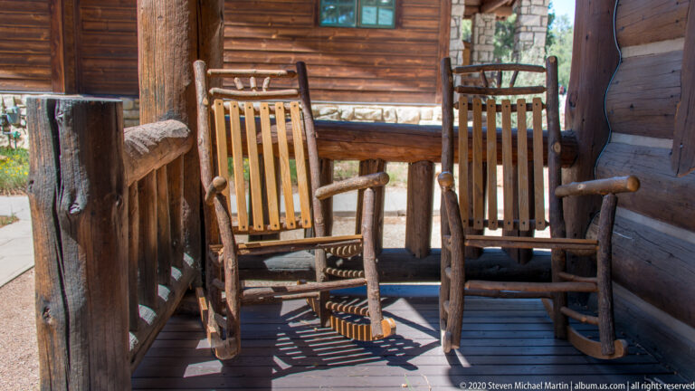 Two slatted wooden rocking chairs sit on a plank cabin porch beside log walls and a stone pier.