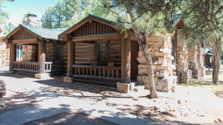 A log cabin with a timber gable and deep porch stands beside a large stone chimney.