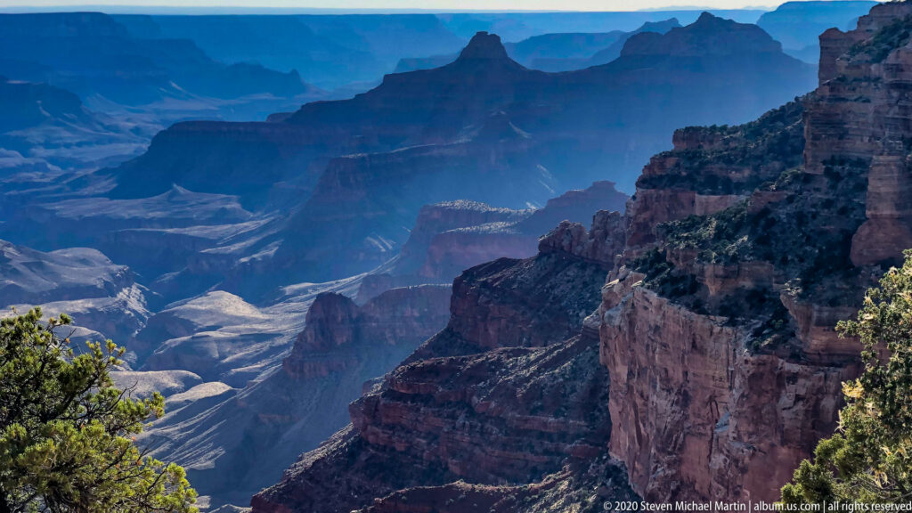 Blue haze softens distant canyon plateaus while a dark cliff fills the right foreground.