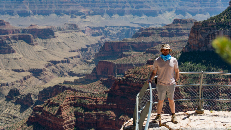 A visitor in a brimmed hat stands beside a metal railing above the canyon.