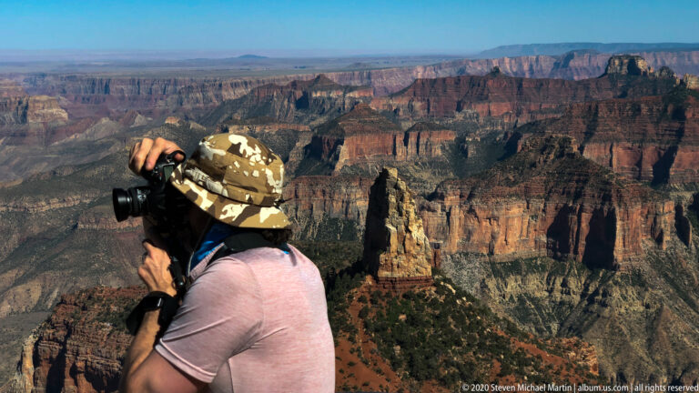 A visitor in a camouflage hat photographs the layered Grand Canyon from a forested rim.