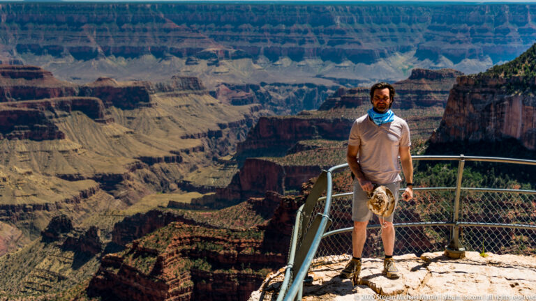 A visitor faces the camera beside a curved rim railing, holding a hat with canyon cliffs behind.