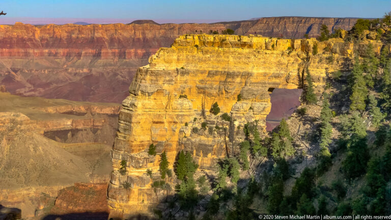 A golden, horizontally layered cliff rises above a wooded slope beside the canyon.