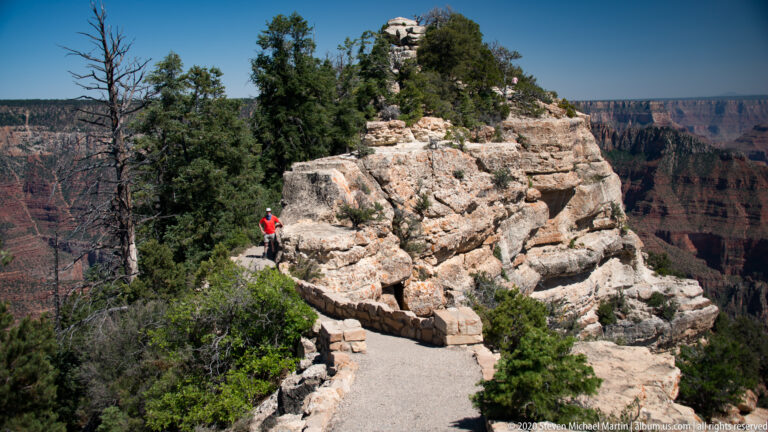 A visitor in red stands on a paved trail beside a rocky outcrop at the canyon rim.