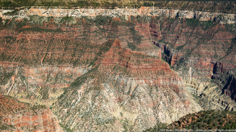 Layered red, cream, and gray canyon walls are cut by a narrow dark ravine.