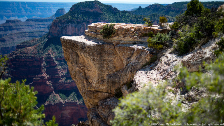 A flat-topped rock ledge with small trees projects above the deep canyon and wooded slope.