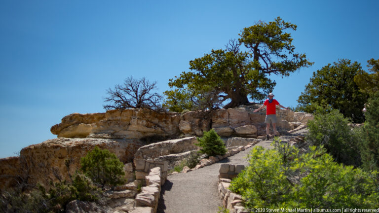 A visitor in red walks up a paved, stone-edged path toward a rocky rim crest.