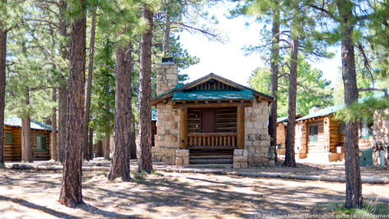 A rustic cabin with a stone gable and chimney, log walls, and a small porch stands among pines.