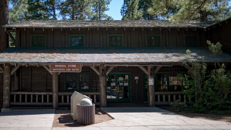 A two-story timber general store has a covered balcony and a sign above its shaded entrance.