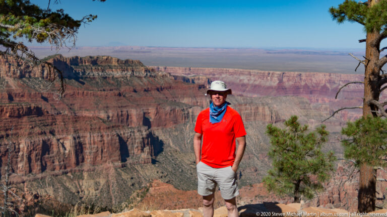 A visitor in a red shirt stands near the canyon rim with layered cliffs behind him.