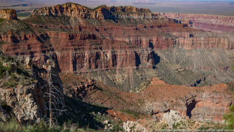 Stratified red and pale cliffs form ridges and ravines beneath a distant plateau.