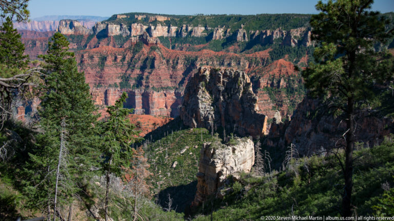 Pale rock towers rise from a wooded gorge beneath broad red and cream canyon walls.
