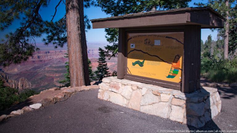 A roofed information kiosk on a stone base stands at a canyon overlook among pines.
