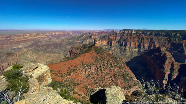 Pale rim boulders and evergreen shrubs frame a canyon promontory and distant layered cliffs.