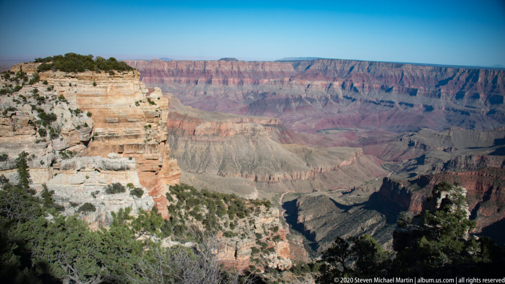 Pale layered cliffs descend at left toward broad red and purple canyon walls beneath a blue sky.
