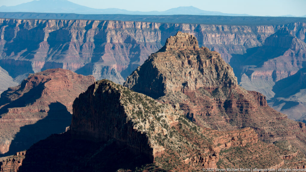 A steep central butte rises above shadowed ravines, backed by layered canyon walls and distant plateaus.
