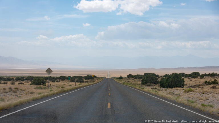 A two-lane road runs straight across a sagebrush basin toward distant mountains beneath clouds.