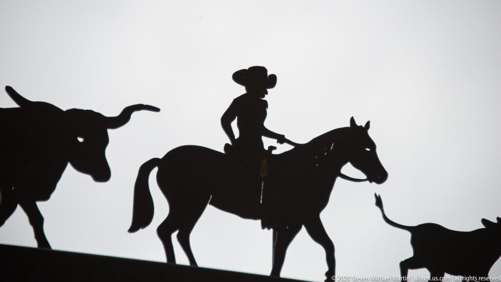 A silhouetted cowboy rides between two horned cattle figures against a bright background.