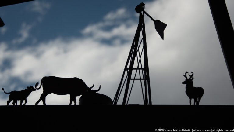 A lattice windmill towers over cattle and deer silhouettes against a blue, clouded sky.