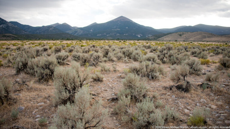 Sagebrush and dry grass cover an open plain below a distant dark mountain range.