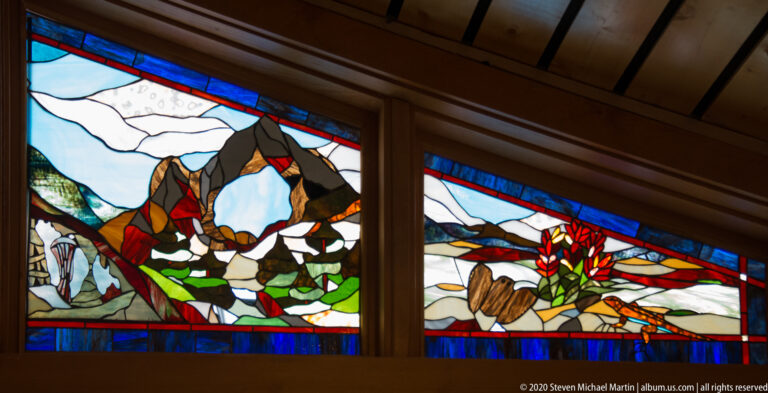Two stained-glass panels show pale peaks and colorful plant and wildlife forms in dark frames.