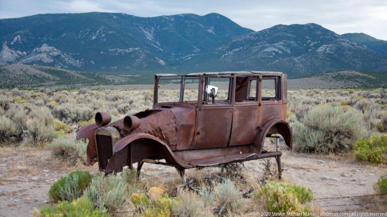 A rusted early automobile with rounded fenders stands in sagebrush before a mountain range.