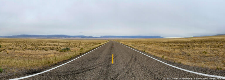 A straight paved road crosses golden shrubland toward a distant low mountain range beneath cloudy sky.