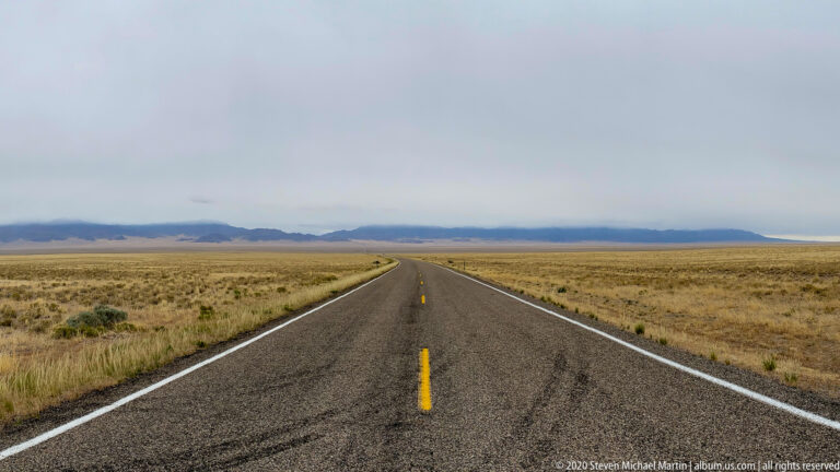A paved road crosses a broad dry plain toward blue-gray mountains under a cloudy sky.