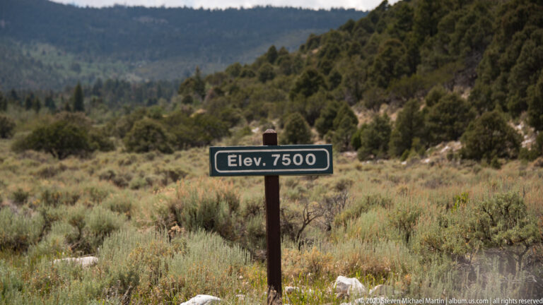 A green “Elev. 7500” marker stands among sagebrush and pale rocks below wooded hills.