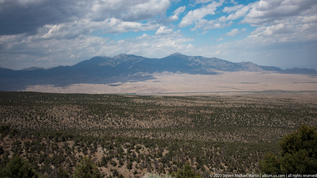 A wide basin and dark forested foothills lead toward a distant mountain range under scattered clouds.