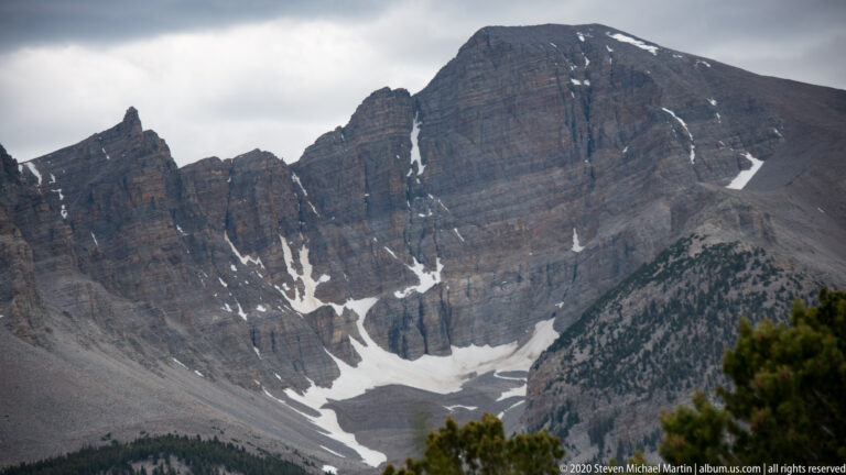 A massive gray rock face rises above a scree basin with patchy snow and a few foreground trees.