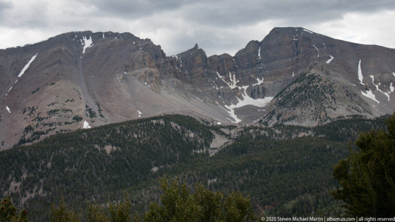 A broad gray mountain arc with snow-filled gullies rises above dark forest beneath clouds.