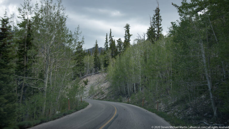 A paved road with a yellow centerline bends through aspens and conifers beside a rocky slope.