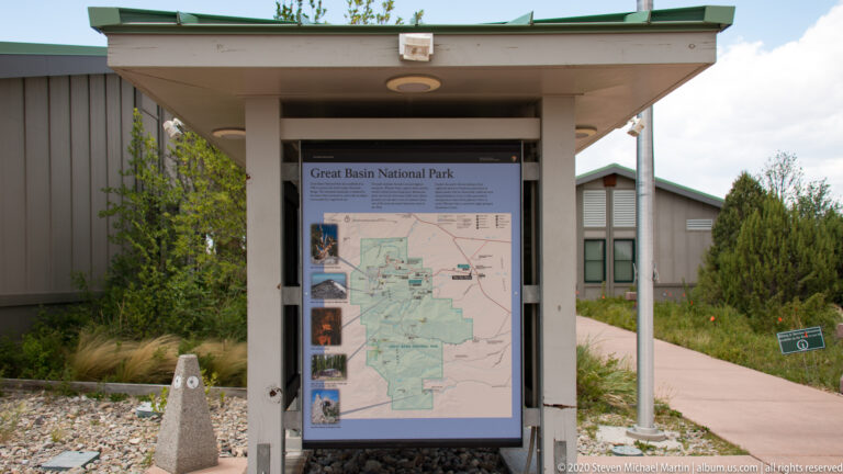 A roofed outdoor kiosk displays a park map beside a visitor-center building and paved walkway.