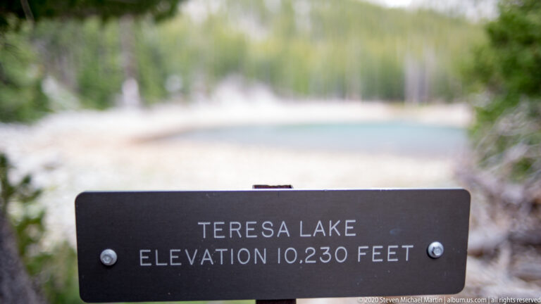 A dark sign reads “Teresa Lake, Elevation 10,230 Feet,” with the lake blurred behind.