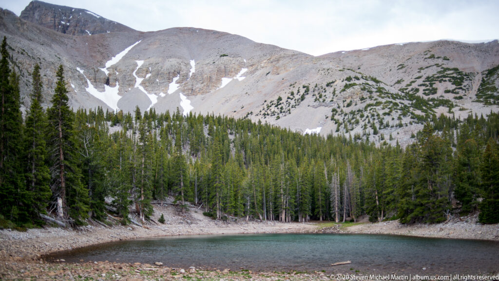 Turquoise Teresa Lake lies below dark conifers and pale snow-patched mountain slopes.