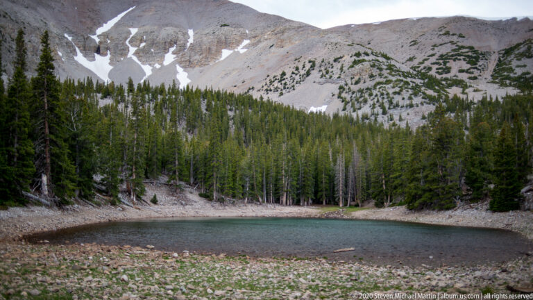 A stony shore leads to blue-green water, with conifers and snow-streaked slopes beyond.