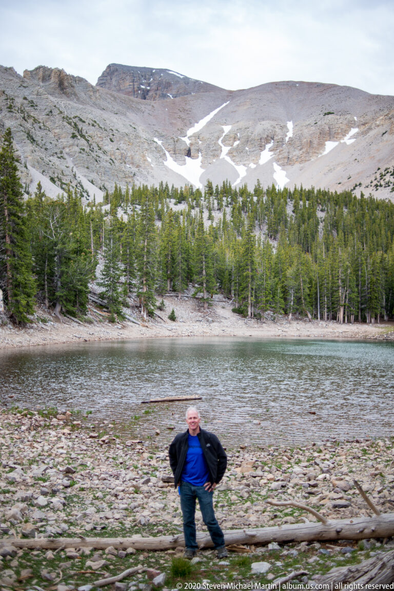 A man stands on Teresa Lake’s rocky shore with forest and snow-patched mountains behind him.
