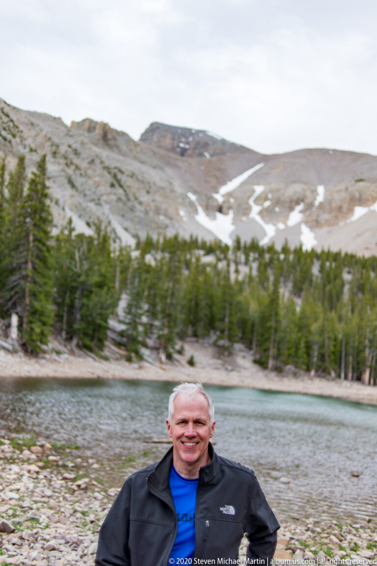 A smiling man in a black jacket and blue shirt stands before a blurred alpine lake.