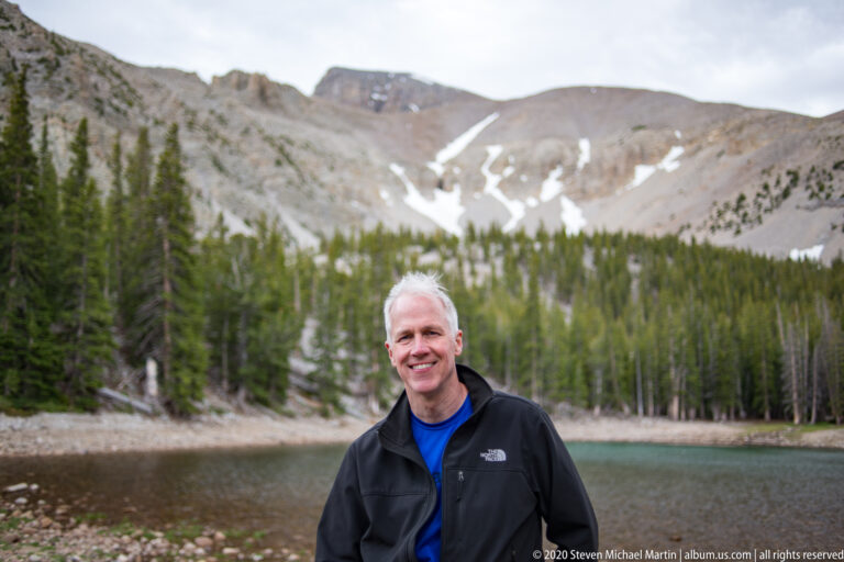 A gray-haired man in a black jacket stands beside Teresa Lake with snowy slopes behind.