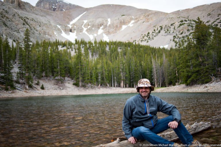 A seated man in a brimmed hat rests on a log at the lake’s stony edge.