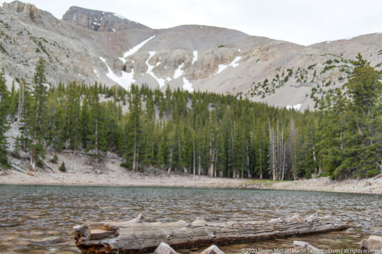 A fallen log crosses the rocky edge of Teresa Lake beneath forest and snow-patched slopes.