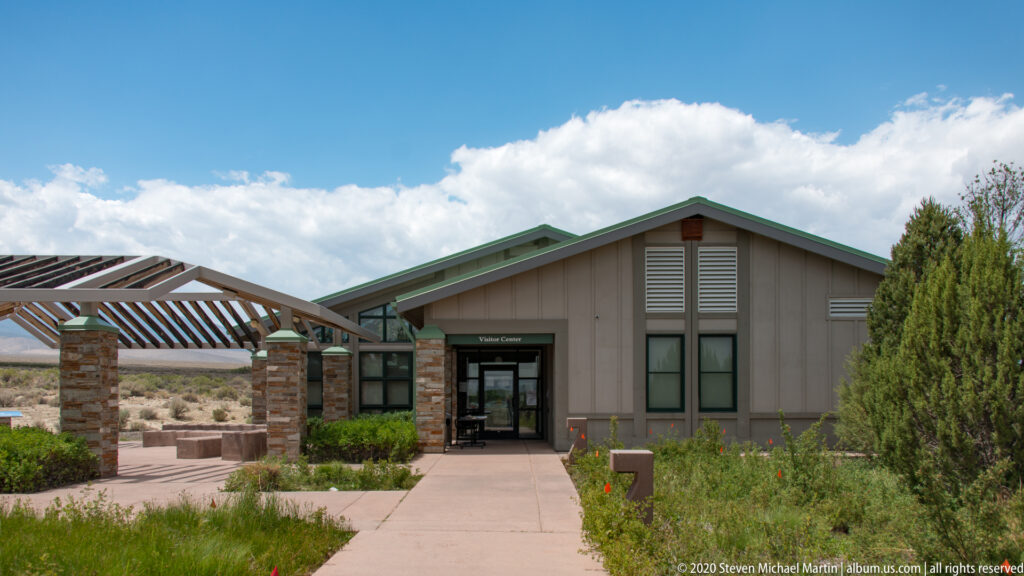 A concrete path leads to a gray visitor center with green roof trim and a timber shade structure.