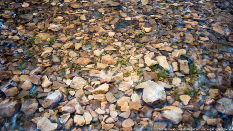 Clear shallow water reveals many multicolored stones across the lake bottom in close view.