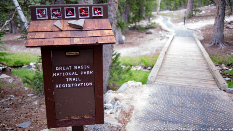 A wooden trail registration box stands beside a boardwalk entrance in the forest.