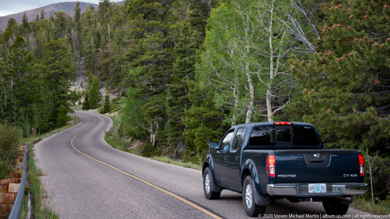 A pickup truck follows a winding paved road through a dense mountain forest.