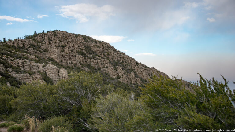 A rocky ridge descends toward sagebrush and scattered pines beneath a hazy blue sky.