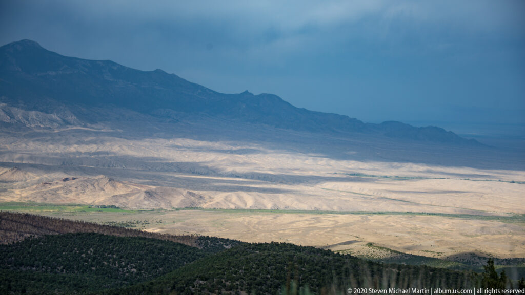 A sunlit basin lies between distant blue mountain ranges, seen above a dark forested slope.