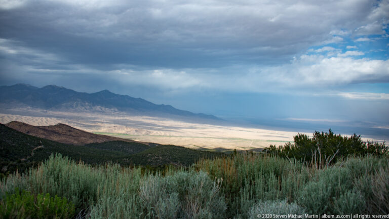 Clouds spread above a broad pale basin and distant range, with shrubs and trees in the foreground.