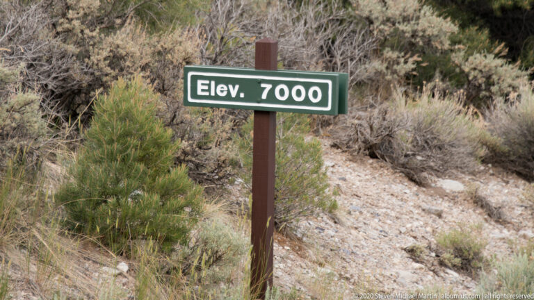 A green “Elev. 7000” sign stands beside a gravel path among dry shrubs and stones.